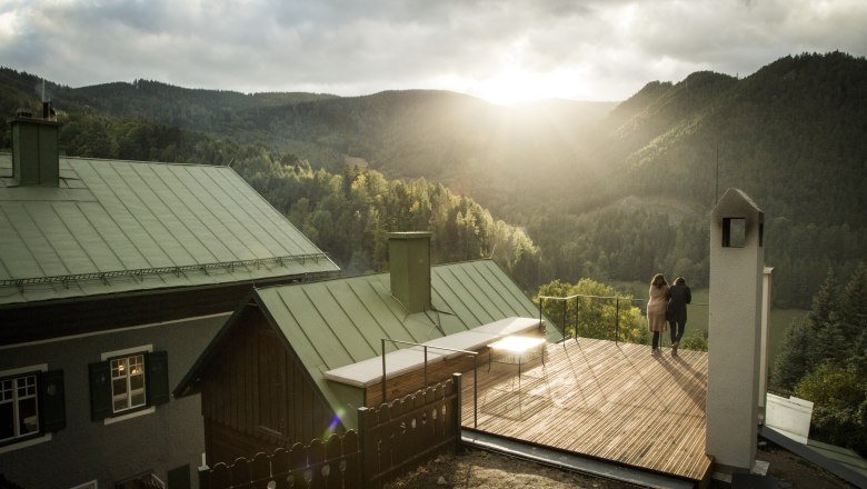 Villa Antoinette, © Matthias Kronfuss Two people stand on a terrace with a view of wooded mountains at sunset.