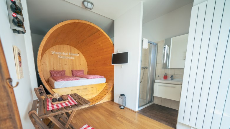 Interior view of a suite with a bed in a large wine barrel, a small table with chairs and a bathroom.