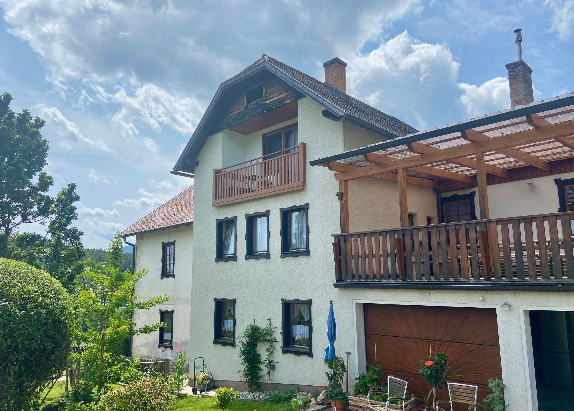A two-story house with balcony and terrace, surrounded by garden and trees, under a cloudy sky.