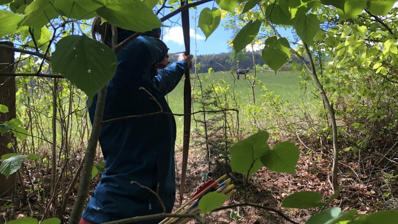 Person shoots with a bow in the forest, surrounded by green leaves.
