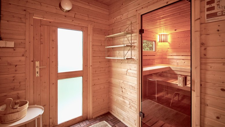 Interior view of a sauna with wooden walls, a glass door and a bench. A wooden bucket stands on a table.