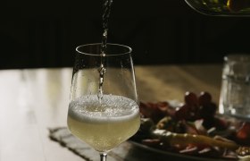 A glass of white wine is poured on a table, with a plate of snacks in the background.