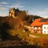 An inn with a red roof and solar panels next to a river, with a castle on a hill in the background.