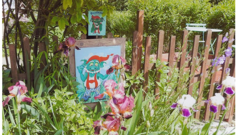 A colorful picture of a green gnome with a red cap in a garden with flowers and a wooden fence.