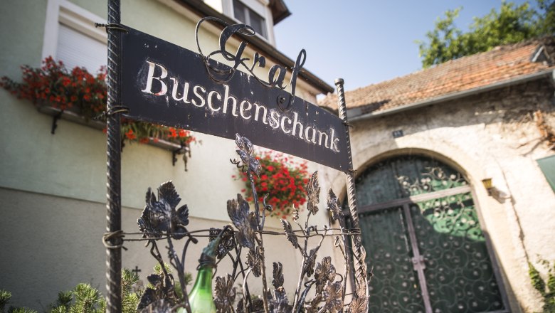 Entrance sign of a Buschenschank (typical tavern) with flowers and gate in the background.