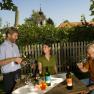 Three people enjoying wine in the garden, with a wooden fence and a church in the background.