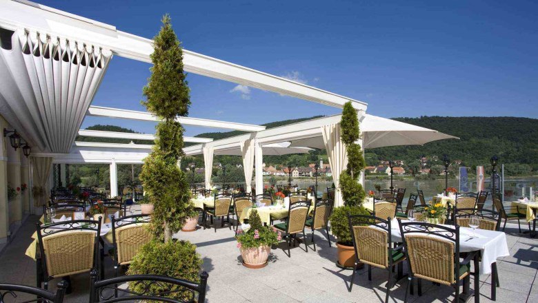 Residence Wachau - Terrace, © Hotel Donauterrasse Terrace of the Wachau residence with tables and chairs, surrounded by plants, with a view of a river and green hills in the background.