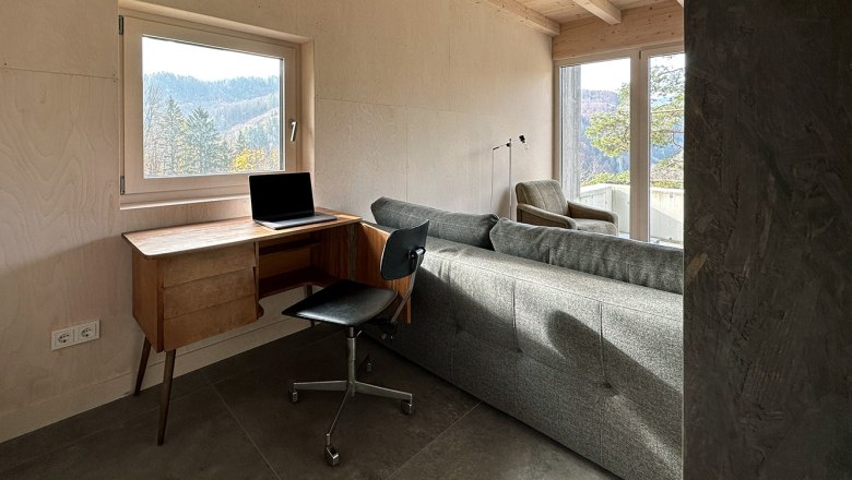 A modern workstation with a laptop on a wooden table, a black chair and a sofa in a room with large windows and a view of the forest.