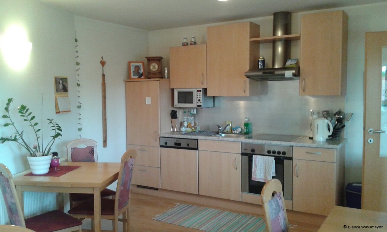 A cozy kitchen with wooden furniture, dining table, chairs, sink, stove and microwave. There are pictures and a calendar on the wall.