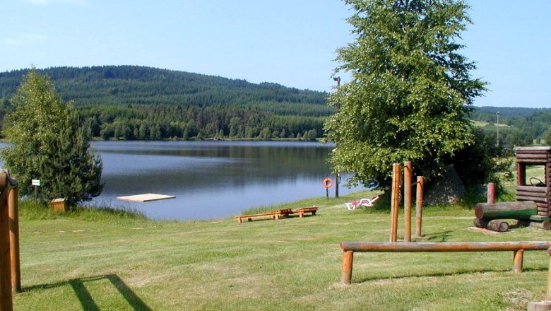 A peaceful lake with a jetty, surrounded by green hills and trees, with a playground in the foreground.