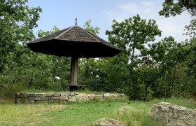 A wooden pavilion with a pointed roof in a green, wooded setting.