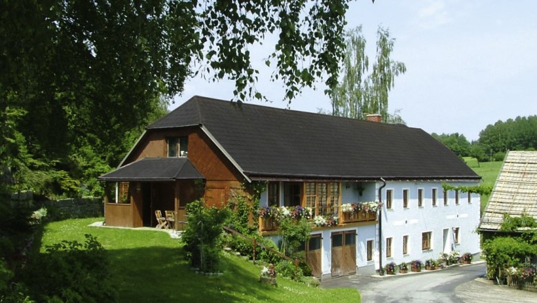 Simandel mill, © Simandelmühle, Johanna Auer A traditional farmhouse with flowers on the balcony, surrounded by green countryside and trees.