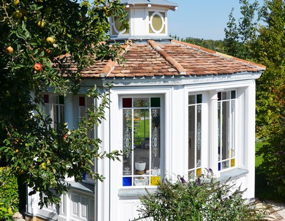 A white pavilion with colorful windows in a garden, surrounded by trees and a fence.