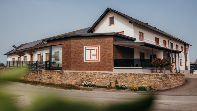 A modern building with the inscription 'Gasthof Thaler', surrounded by a stone wall and plants.