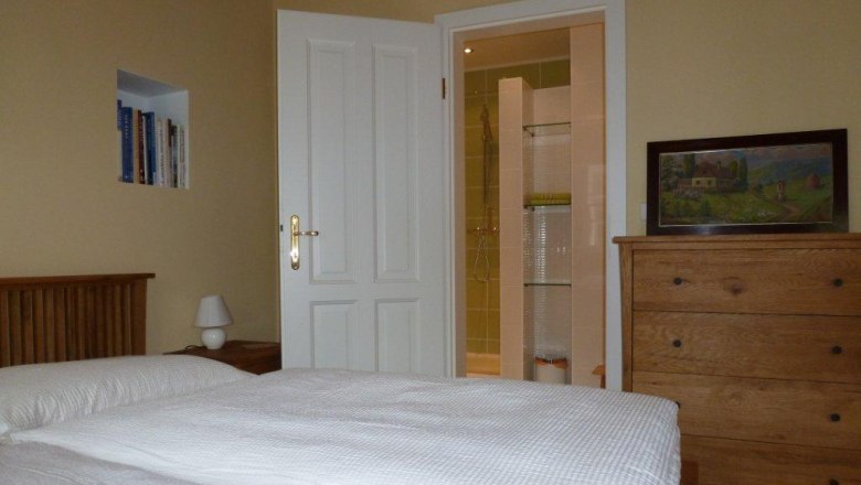 A cozy double room with bed, chest of drawers, bookshelf and open bathroom.