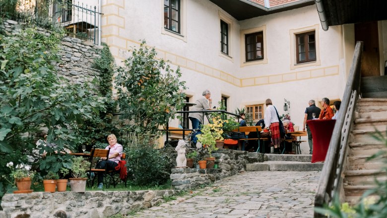 An inner courtyard with people sitting and standing at tables, surrounded by plants and a stone building.