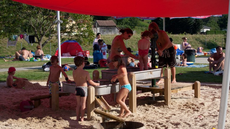 Children play in the sand under a red awning in Wilhelmsburg Parkbad.