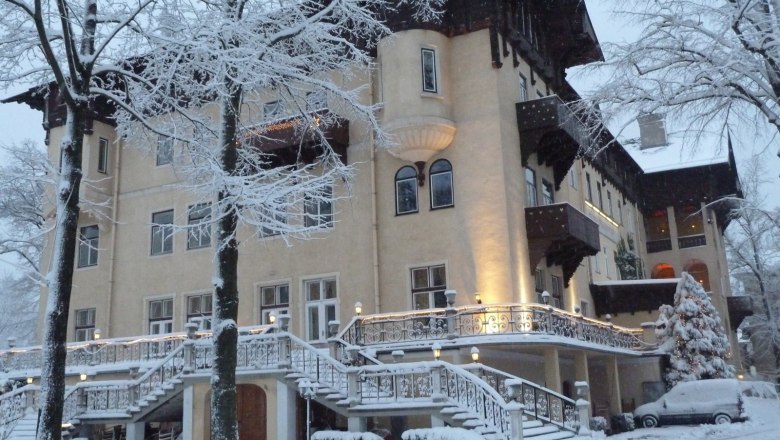 A large, elegant hotel in winter, surrounded by snow-covered trees and a snow-covered car.