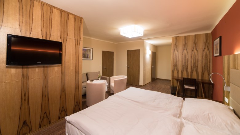 Modern hotel room with double bed, TV, seating area and wooden furniture.
