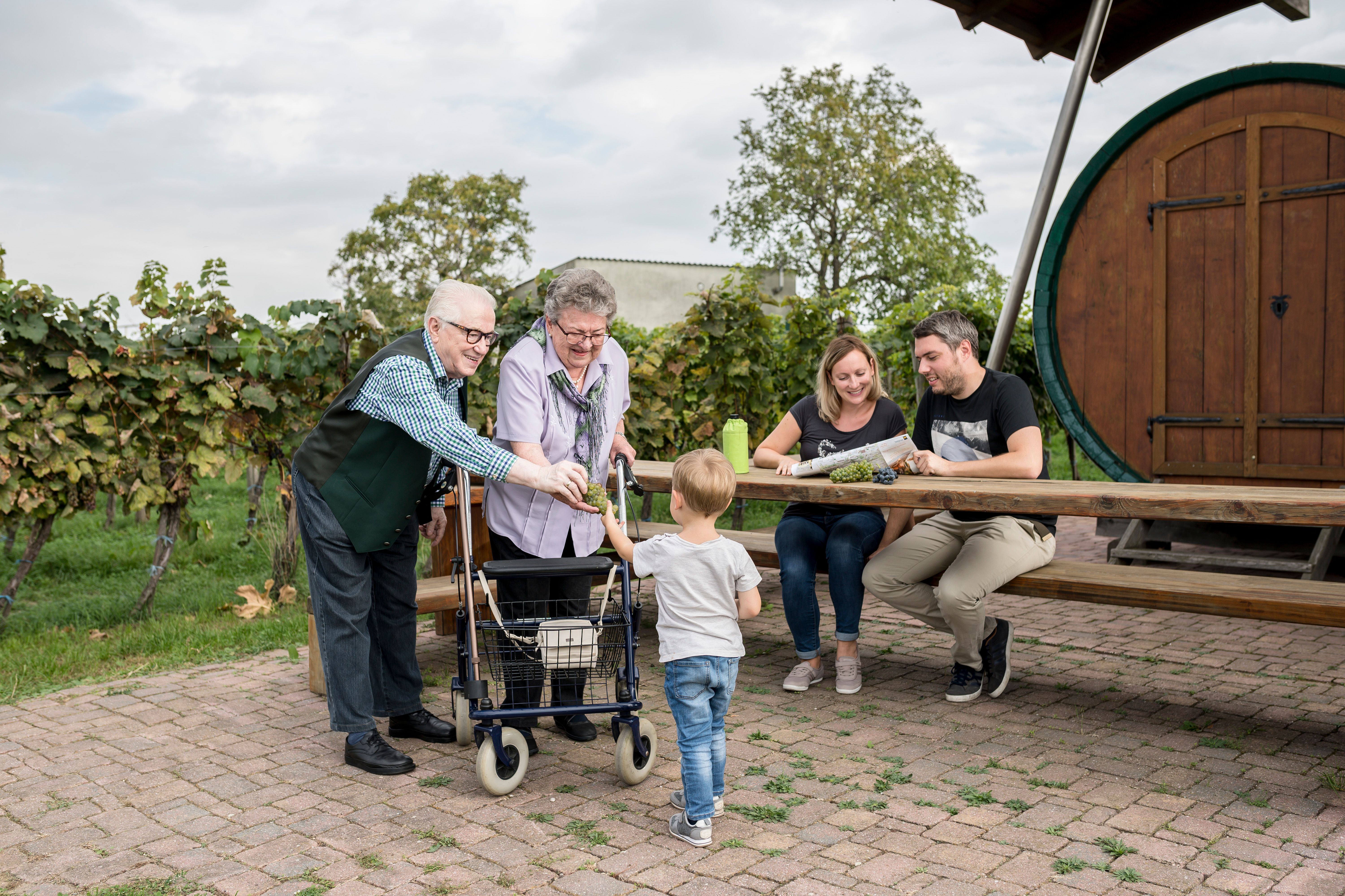 A family sits at a wooden table outside, surrounded by vines. An elderly couple is giving grapes to a child.