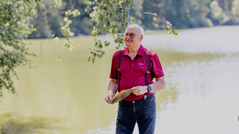 An elderly man in a red shirt stands smiling on the shore of a lake, holding a piece of fish skin in his hands.