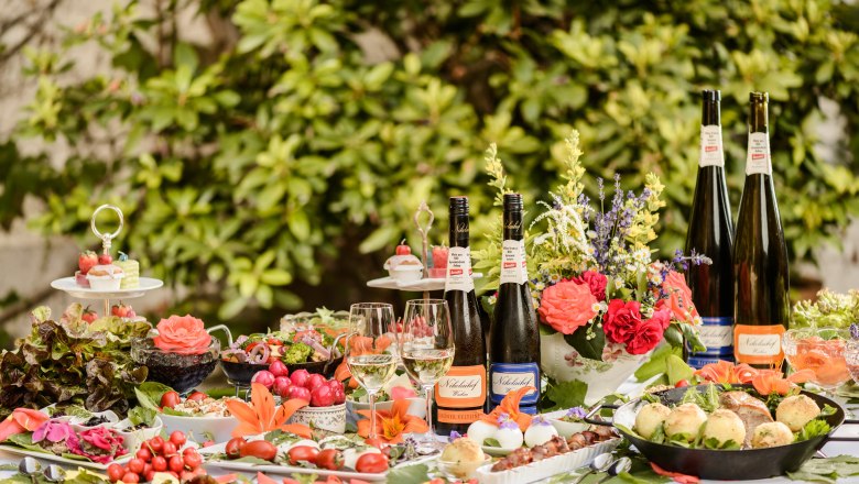 A richly laid outdoor table with bottles of wine, glasses, flowers and various dishes.