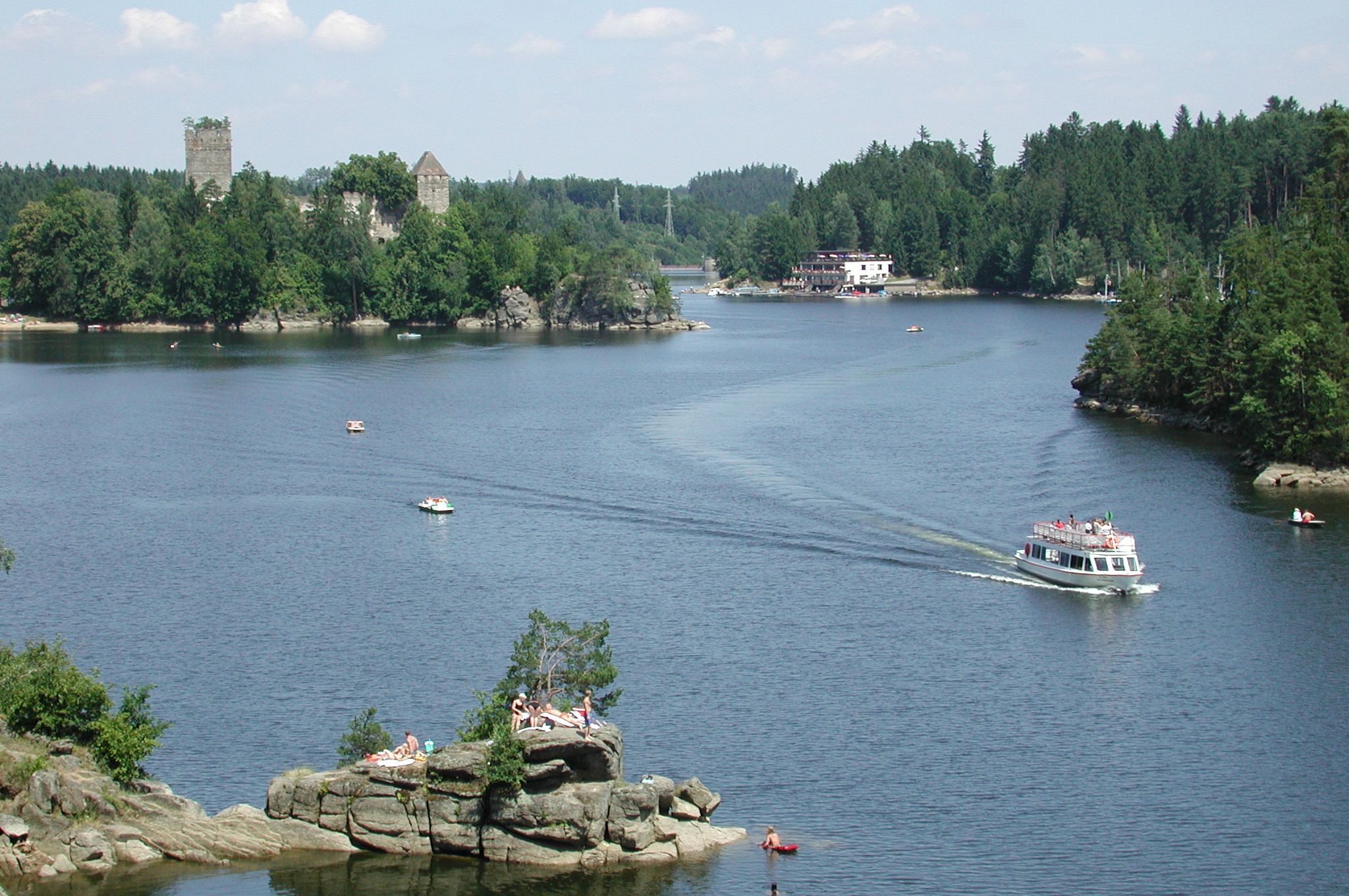 A lake with boats, a castle in the background and people on rocks.