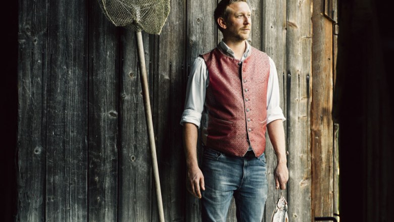 Man in red vest standing in front of wooden wall with fishing net and fish.