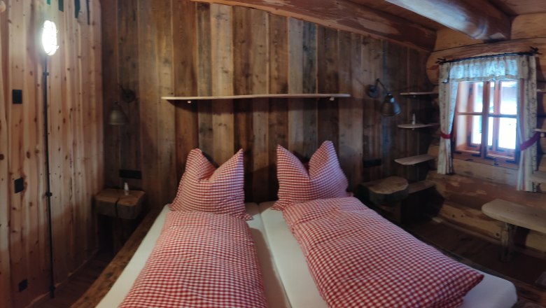 Rustic room with wooden walls, bed with red and white checkered pillows, window with curtains, wooden door and shelves.