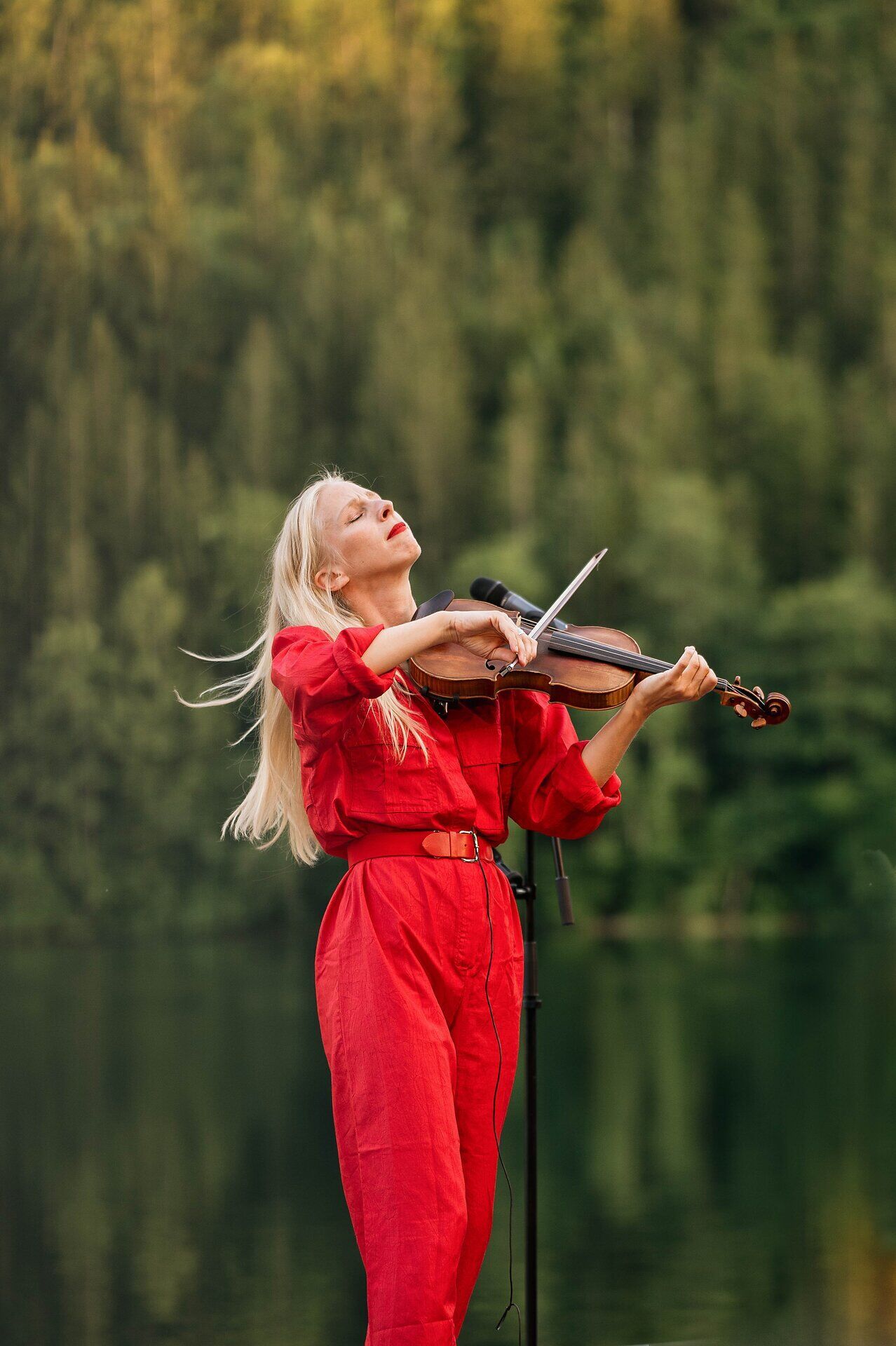 Geigerin in rotem Overall spielt im Freien vor einem ruhigen See, die Augen geschlossen, umgeben von dichtem, grünem Wald.