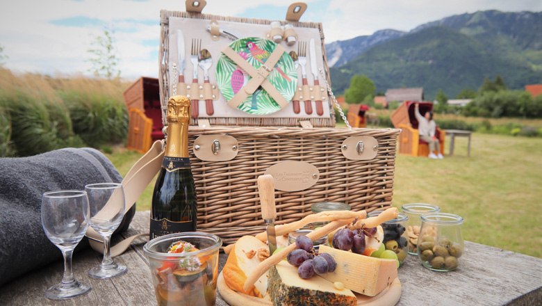 Picnic basket with cheese, grapes and bottle of champagne on a table in a rural setting with mountains in the background.