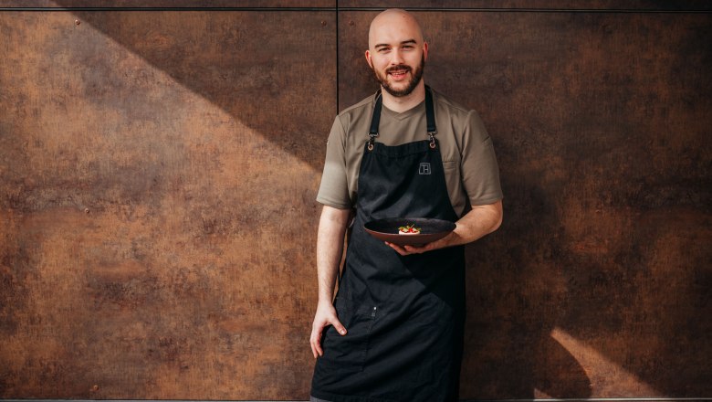 A man in a black apron holds a plate of food in front of a brown wall.