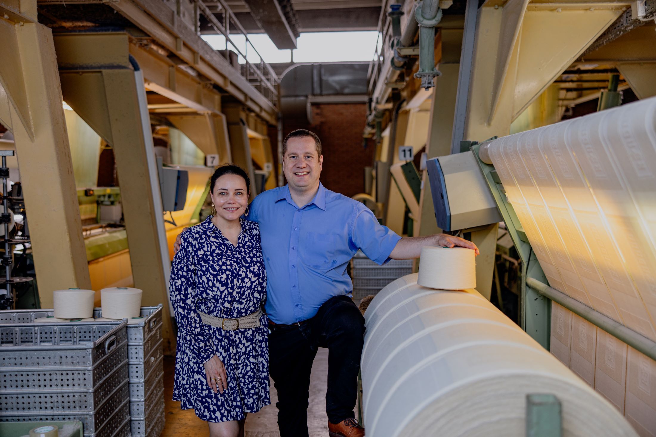 Two people stand next to machines and rolls of fabric in a textile factory.