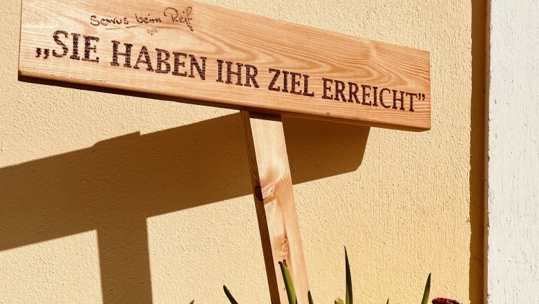 Wooden sign with the inscription 'You have reached your destination' in front of a yellow wall surrounded by flowers.