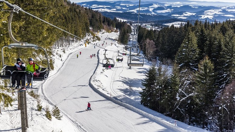 Ski area with chair lift and skiers, surrounded by snow-covered trees and hills.
