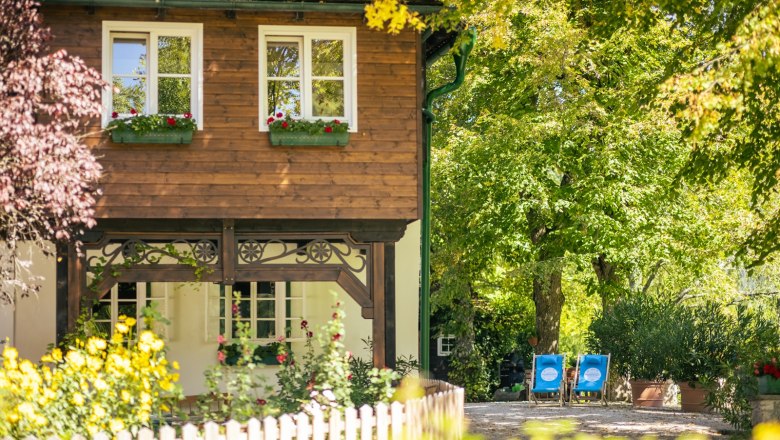 An idyllic country house with wooden elements, surrounded by flowering gardens and trees. There are two blue sun loungers in the garden.