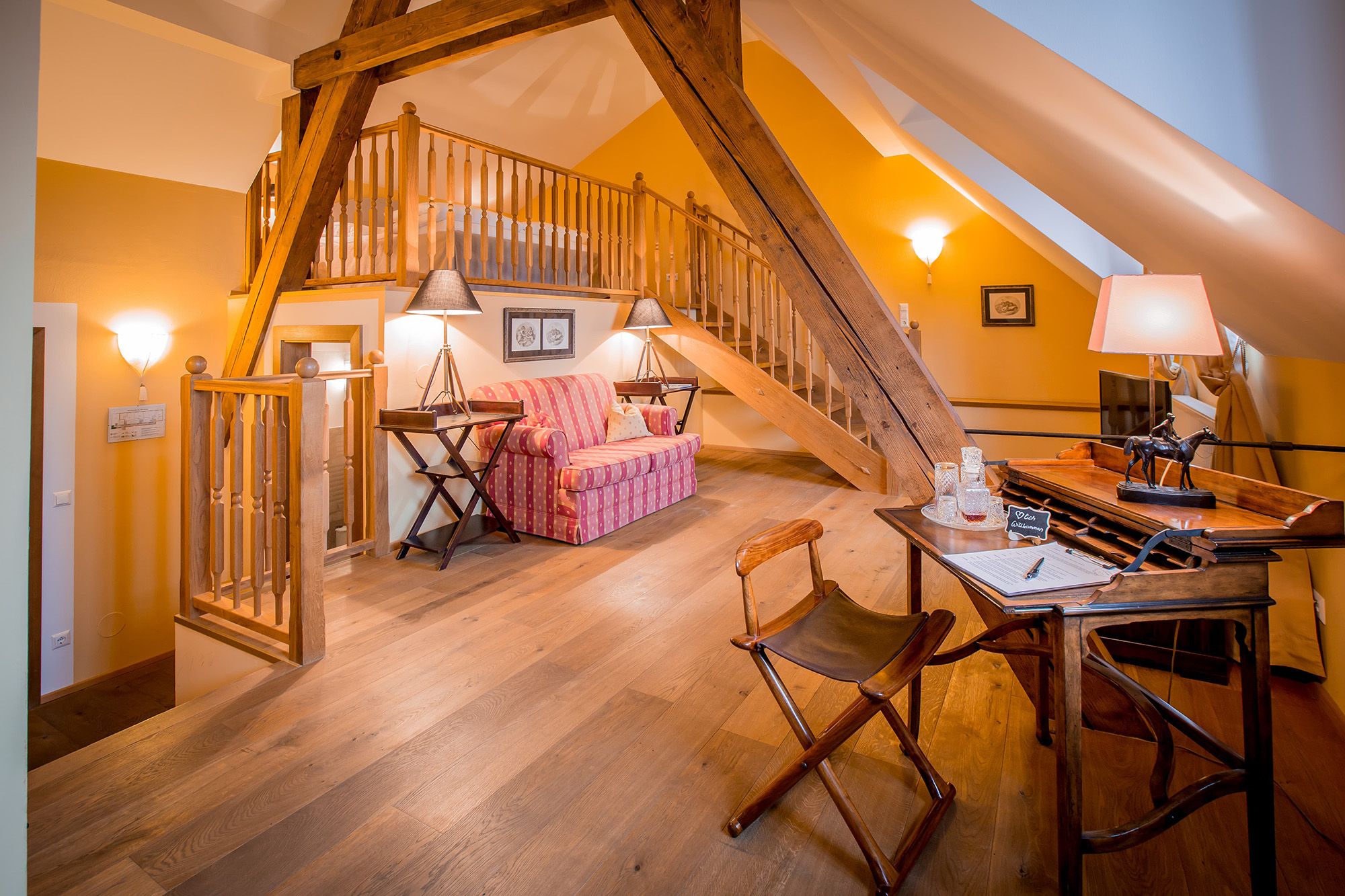 Cozy attic suite with wooden beams, sofa, desk and stairs to a sleeping area.
