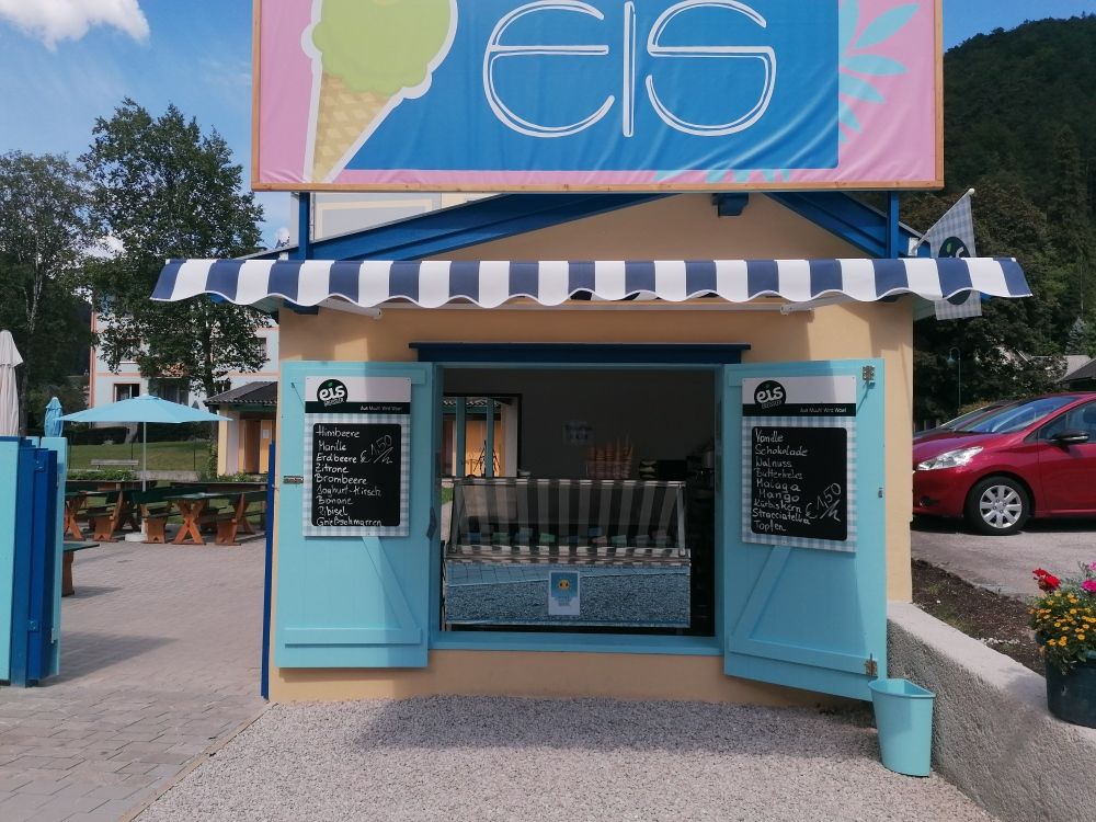 A small ice cream stand with blue shutters and an awning, surrounded by trees and cars.