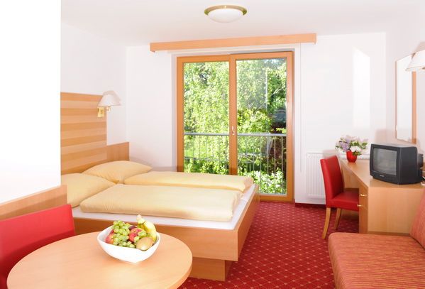 Bright hotel room with double bed, red carpet, table with fruit bowl and TV, view of green trees through a large window.