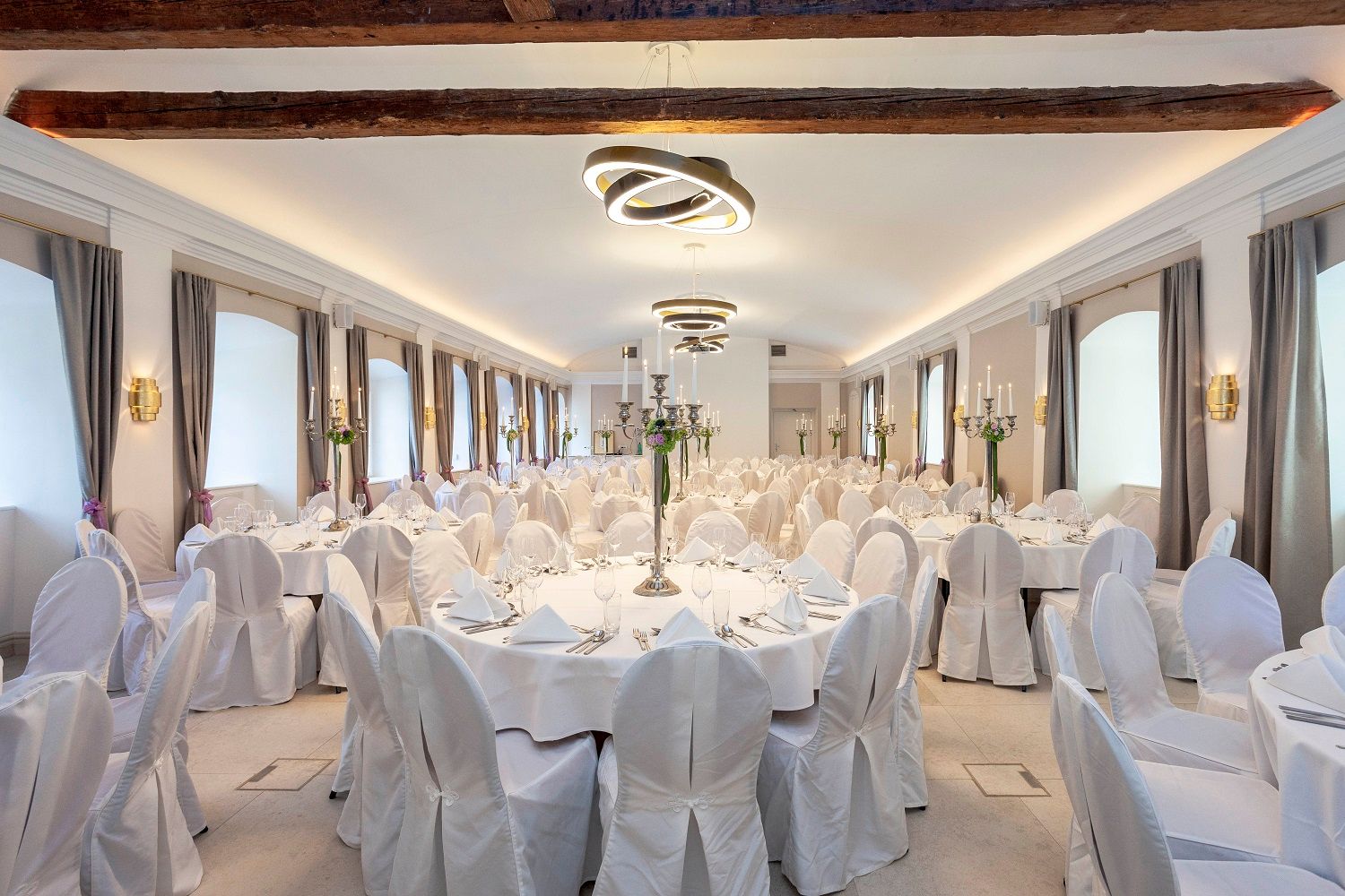 A festively decorated hall with round tables, white tablecloths and chair covers, candlesticks and modern lighting.