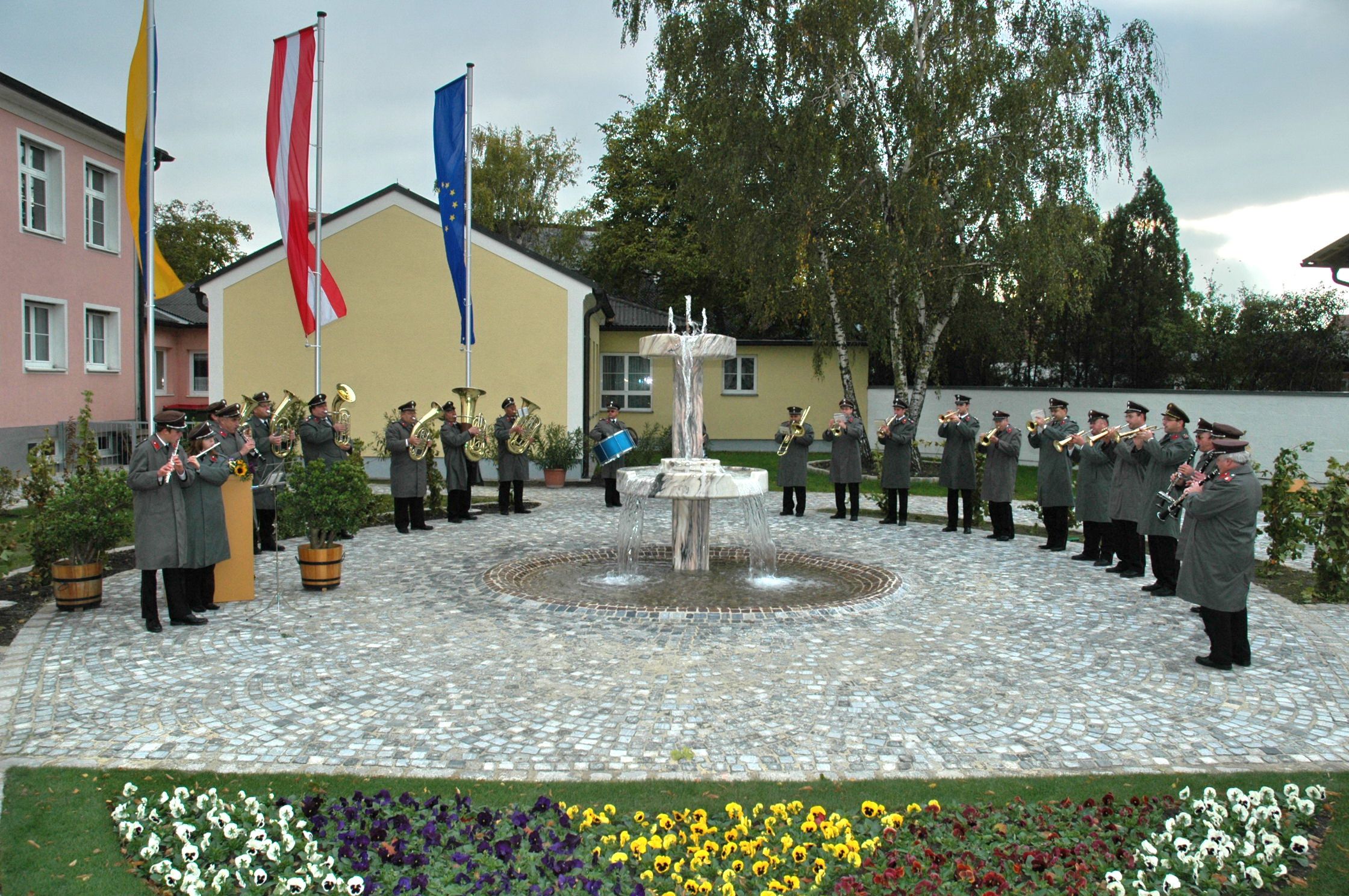 A brass band plays outside in front of a fountain, surrounded by flowers and flags.