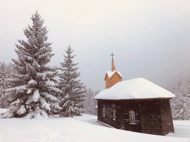 A small church in the snow with fir trees in the background.