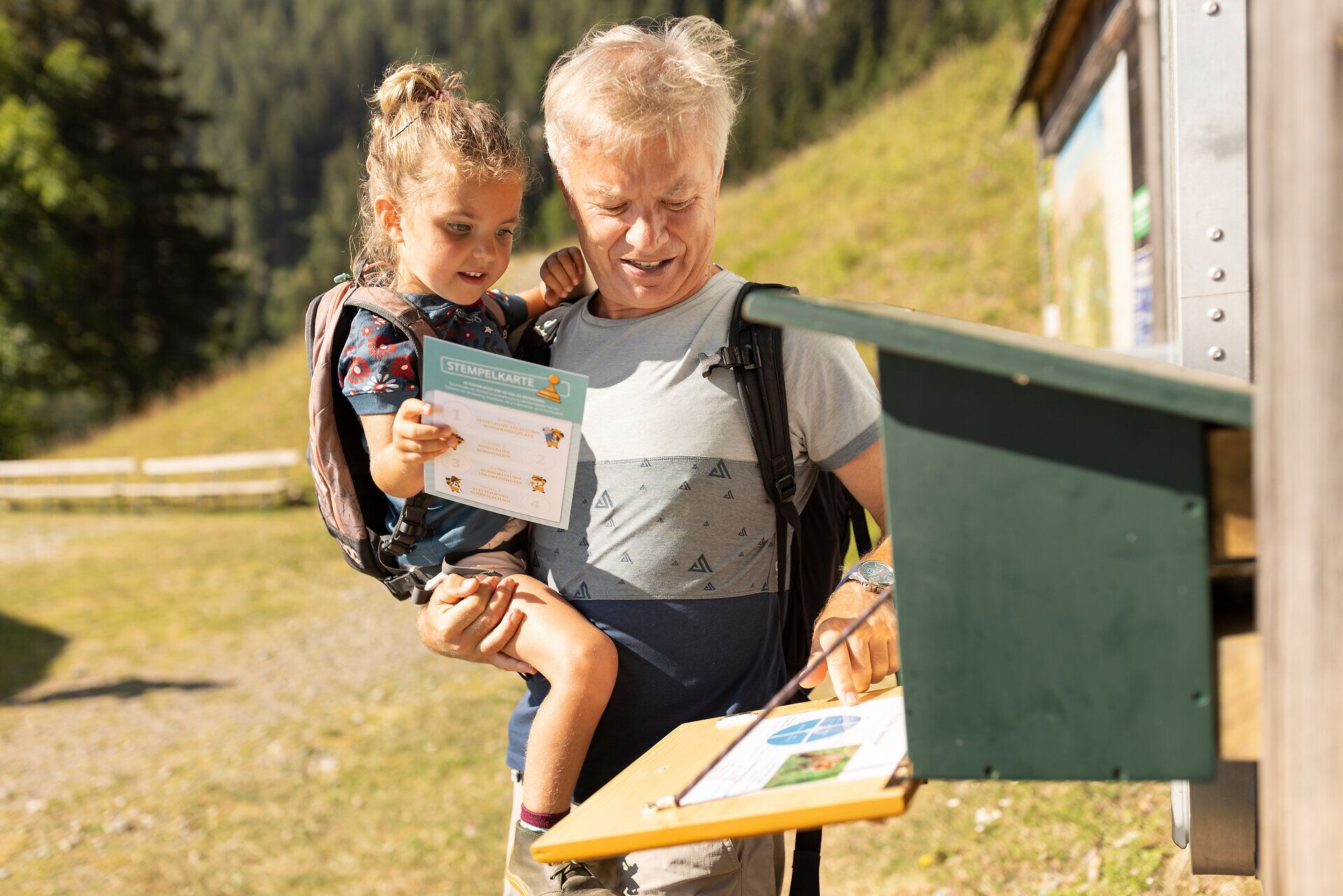 A cheerful grandfather holds his granddaughter in his arms as they stand together at an information booth. The picturesque surroundings of the Vienna Alps invite visitors to enjoy unforgettable hiking adventures that bring families together and allow them to experience the beauty of nature.
