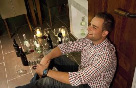 A man sits smiling on the floor, surrounded by bottles of wine and glasses.