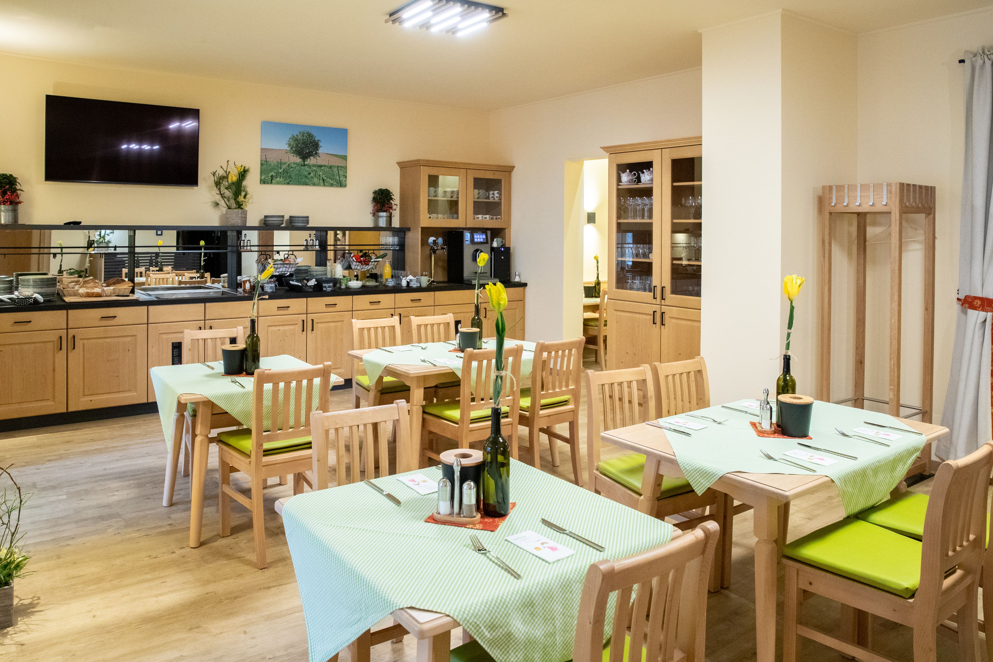 A cozy breakfast room with wooden furniture, laid tables and a buffet area. Pictures hang on the walls and plants decorate the room.