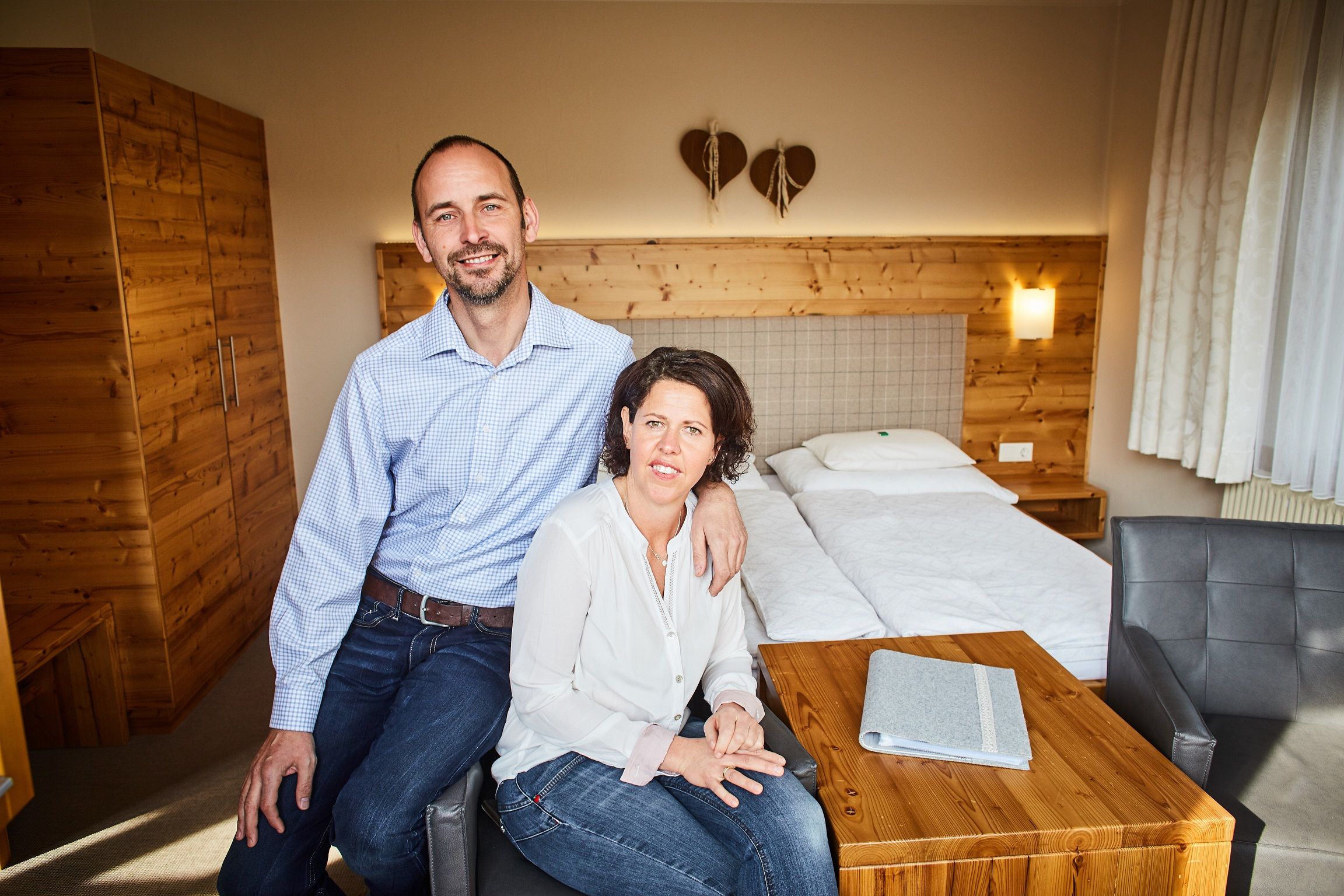 A couple is sitting in a cozy room with wooden furniture and a double bed.