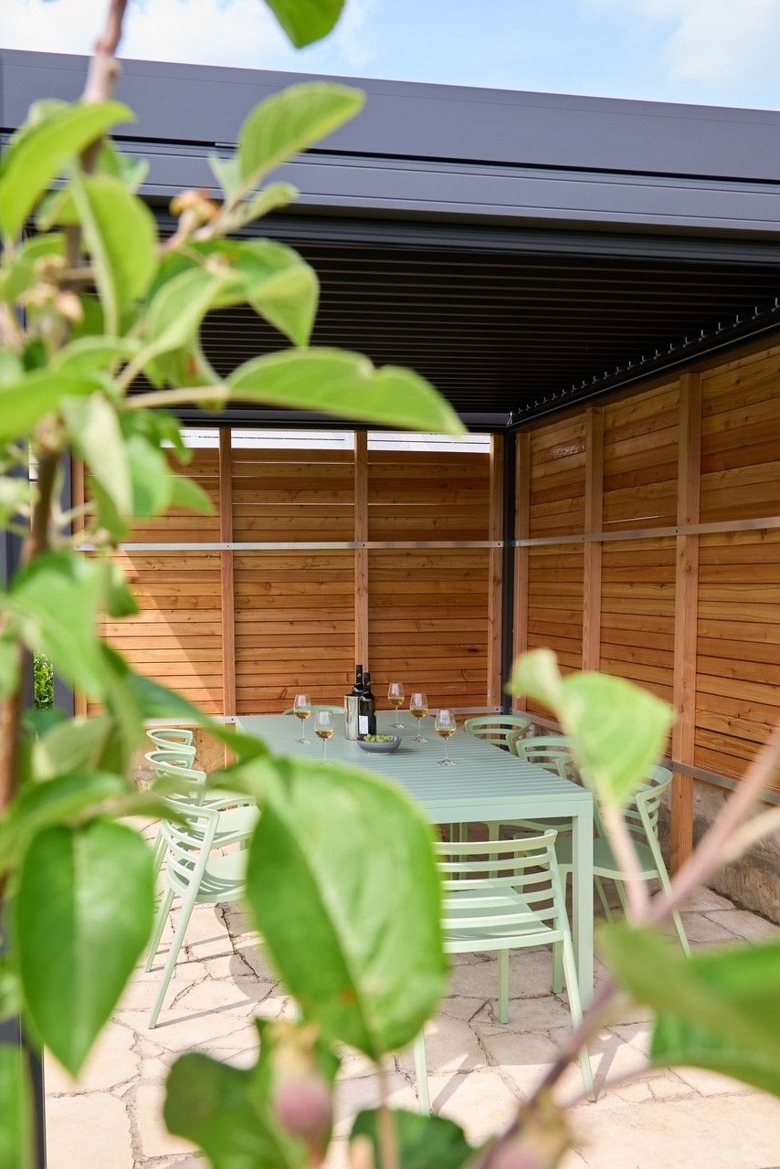 Covered terrace with table and chairs, surrounded by wood paneling and plants in the foreground.