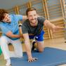A man does an exercise on a mat, supported by a woman in a fitness studio.