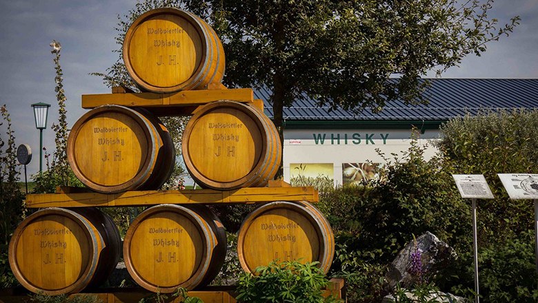 Wooden barrels in the open with the inscription 'Waldviertler Whisky J.H.' in front of a building with a 'Whisky' sign.