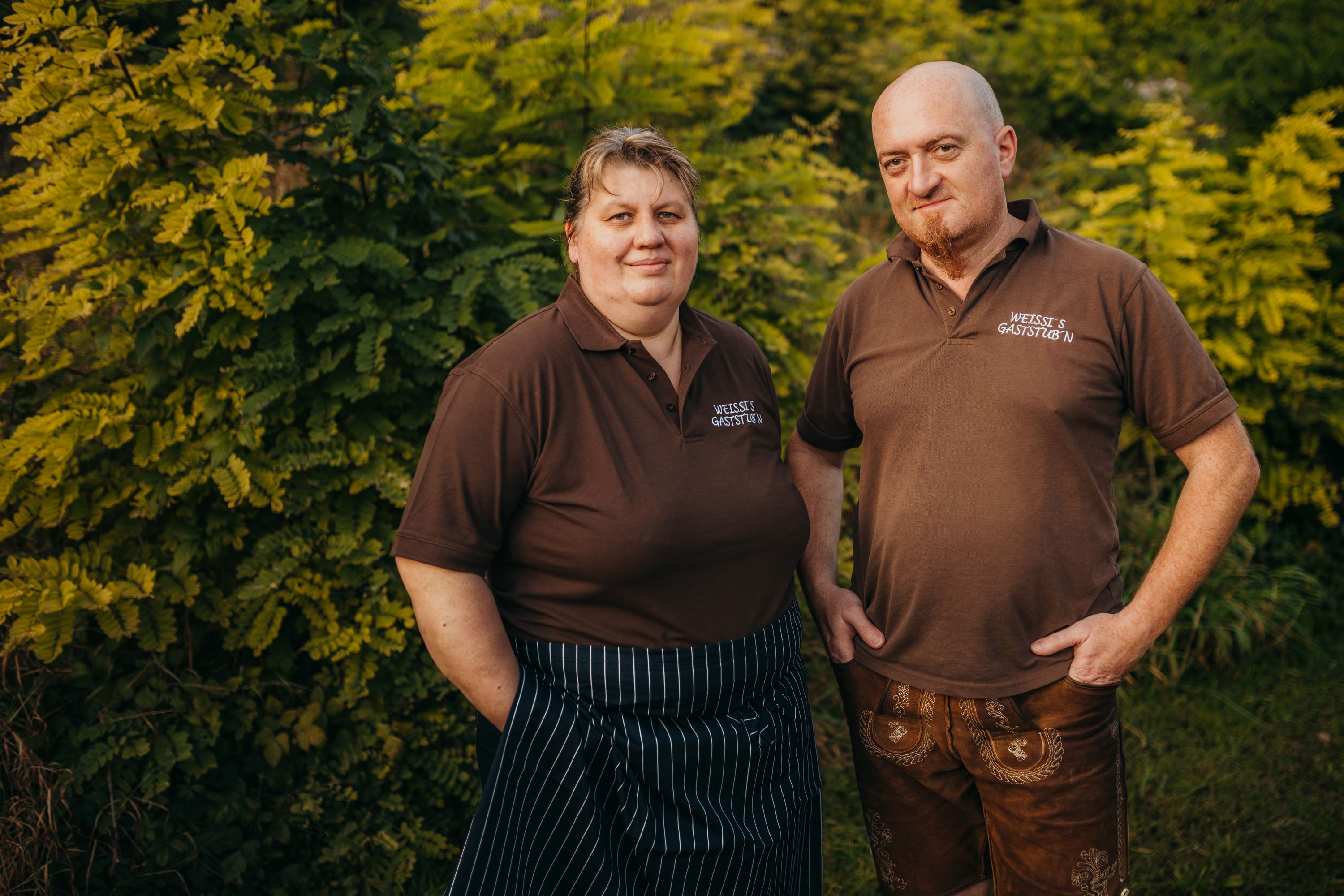 Two people in brown polo shirts in front of green bushes.